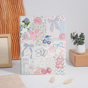 May include: A decorative notebook with a patchwork design featuring floral patterns, bows, cherries, playing cards, and a gummy bear. The color palette includes pink, blue, and white, creating a whimsical and feminine aesthetic.