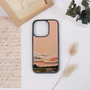 May include: A dark-rimmed smartphone case featuring a printed landscape design. The image displays a textured, impasto-style painting of a sunset sky with peach, pink, and cream clouds above a dark green field and a winding path at the bottom.