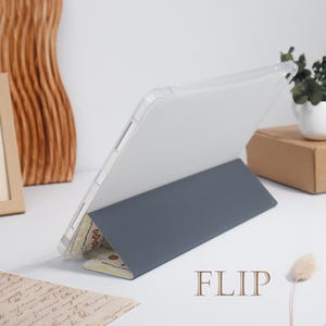 K&ouml;nnte beinhalten: Eine transparente Tablet-H&uuml;lle mit grauem Klappdeckel. Der Deckel ist hochgeklappt und st&uuml;tzt das Tablet in einem Winkel. Der untere Rand des Covers hat ein Muster. Das Wort "FLIP" ist braun gedruckt.