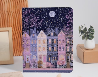 May include: A rectangular notebook featuring a nighttime illustration of a row of colorful townhouses under a starry sky with a full moon. The buildings are rendered in shades of purple, blue, and cream, with glowing windows and flowering trees.