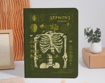 May include: A dark olive green notebook featuring a white skeletal rib cage illustration, botanical elements, and text that reads "It's either the GROWING IS SLOW or it CUTS TO THE BONE" along with smaller phrases and anatomical diagrams.