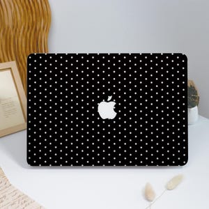 May include: Black laptop case with a white polka dot pattern. The Apple logo is centered on the case. The case is on a white surface with a wooden frame and decorative elements in the background.