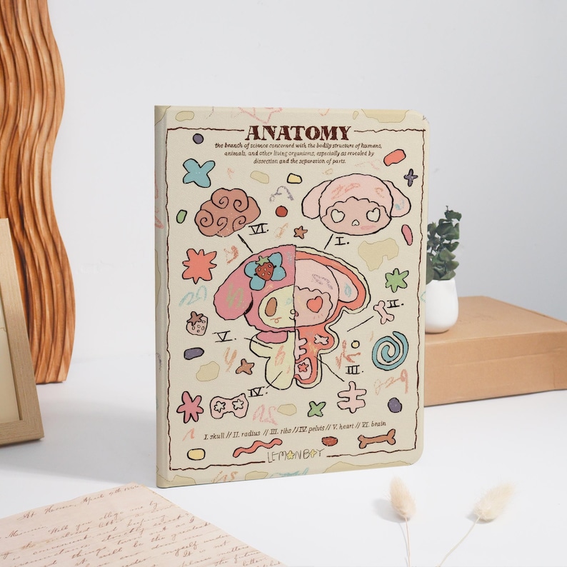 K&ouml;nnte beinhalten: Ein beigefarbenes Notizbuch mit dem Wort "ANATOMY" oben, das eine Cartoon-Illustration eines Charakters zeigt, der in zwei H&auml;lften geteilt ist, um innere Organe zu zeigen. Das Notizbuch hat verschiedene bunte Kritzeleien und anatomische Diagramme. Der Text "LEMONBOY" steht unten.