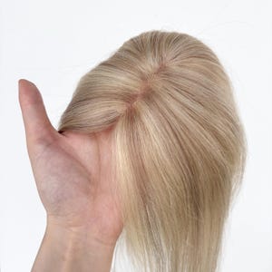Puede incluir: Una pieza de cabello rubio con una base marrón claro. La pieza de cabello se sostiene en una mano sobre un fondo blanco. El cabello es liso y parece ser cabello humano.