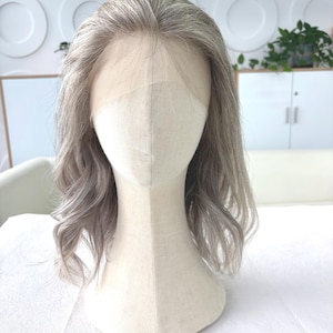 May include: A light gray wig with wavy, shoulder-length hair is displayed on a mannequin head. The wig features a lace front for a natural hairline. The mannequin is set against a white background, with a glimpse of a white cabinet and greenery.