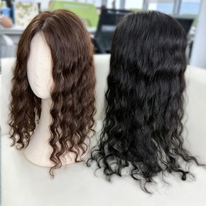 Puede incluir: Dos pelucas de longitud hasta los hombros con textura ondulada. Una peluca es de color marrón medio y se muestra sobre una cabeza de maniquí, mientras que la otra es de color negro sólido y está colocada al lado. Ambas tienen raya central.