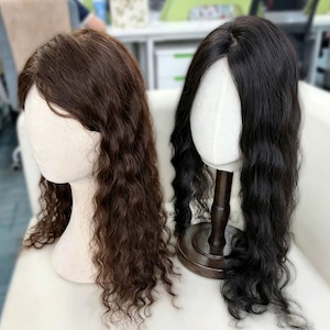 Puede incluir: Dos pelucas largas y onduladas sobre cabezas de maniquí. La peluca de la izquierda es de color marrón medio, mientras que la de la derecha es de color negro intenso. Ambas tienen una raya central y llegan por debajo de los hombros.