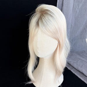 May include: A shoulder-length wig featuring straight, platinum blonde hair with dark roots. The hair is styled with a center part and soft, subtle waves that frame the face. The wig is displayed on a smooth, beige, featureless mannequin head.