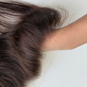 May include: Close-up of a dark brown lace front wig. The wig features long, wavy hair with a natural-looking hairline. The lace is a light brown colour, designed to blend with the skin. The hair is a rich, dark brown colour.