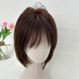 以下が含まれることがあります： 短い、ダークブラウンのウィッグで、カットと前髪が特徴です。マネキンの頭にスタイルされており、髪の自然な質感と色を示しています。髪は豊かで温かみのある茶色で、前髪は顔を縁取るようにカットされています。