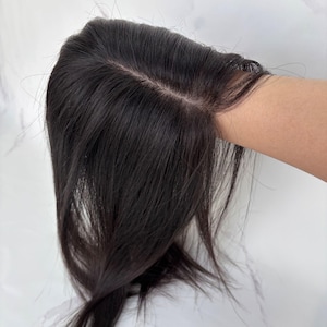 Puede incluir: Una pieza de cabello castaño oscuro con una raya de aspecto natural. El cabello es liso y parece ser cabello humano. La pieza capilar está sujeta por una mano, mostrando la textura y el color del cabello.