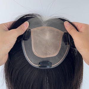 May include: A dark brown hair topper with a sheer base and clips. The hairpiece is held up by two hands, showcasing the natural-looking hair and the attachment clips. The hair topper is designed to add volume and coverage.