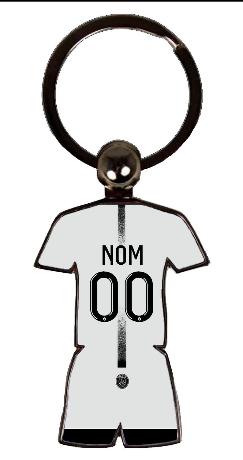 May include: A white jersey-shaped keychain with the text "NOM" above the number "00" in black. The keychain has a black ring and a silver-colored ball at the top. The team logo is at the bottom.
