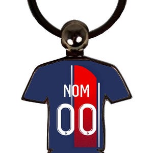 May include: A navy blue keychain in the shape of a soccer jersey. The jersey has a red stripe down the center, the number "00" in white, and the word "NOM" in white. The keychain has a black ring and a small black ball at the top.