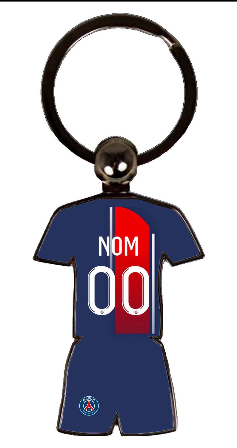 May include: A navy blue keychain in the shape of a soccer jersey. The jersey has a red stripe down the center, the number "00" in white, and the word "NOM" in white. The keychain has a black ring and a small black ball at the top.