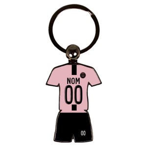 May include: A pink and black soccer jersey keychain with the text "NOM" and the number "00". The keychain has a black ring and a small silver ball at the top. The jersey is a light pink color.