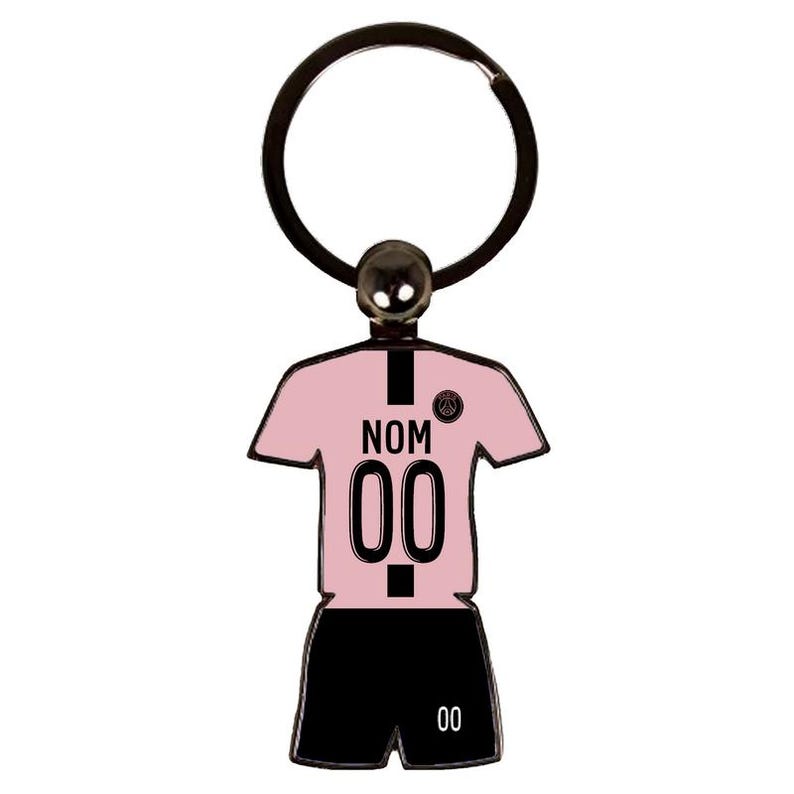 May include: A pink and black soccer jersey keychain with the text "NOM" and the number "00". The keychain has a black ring and a small silver ball at the top. The jersey is a light pink color.
