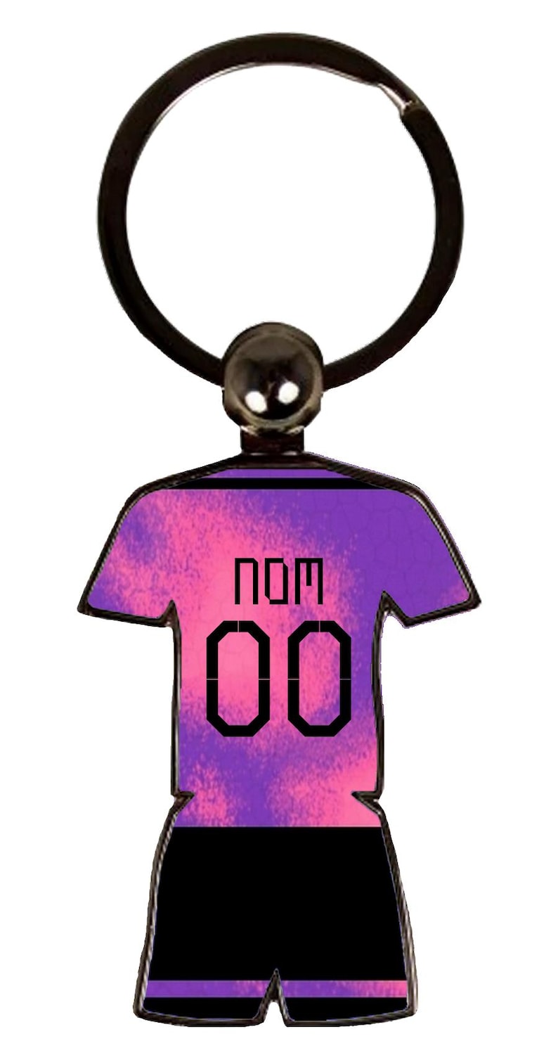 May include: A purple and pink jersey-shaped keychain with the word "NOM" and the number "00" printed on the front. The keychain has a black ring and a small, round, silver-colored attachment.