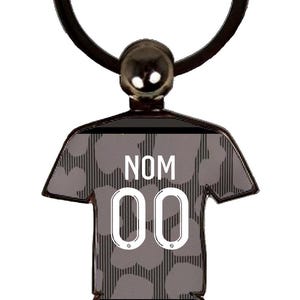 May include: A black and gray keychain in the shape of a soccer jersey and shorts. The jersey has the text "NOM" above the number "00". The keychain has a silver-colored ring and a small ball at the top.