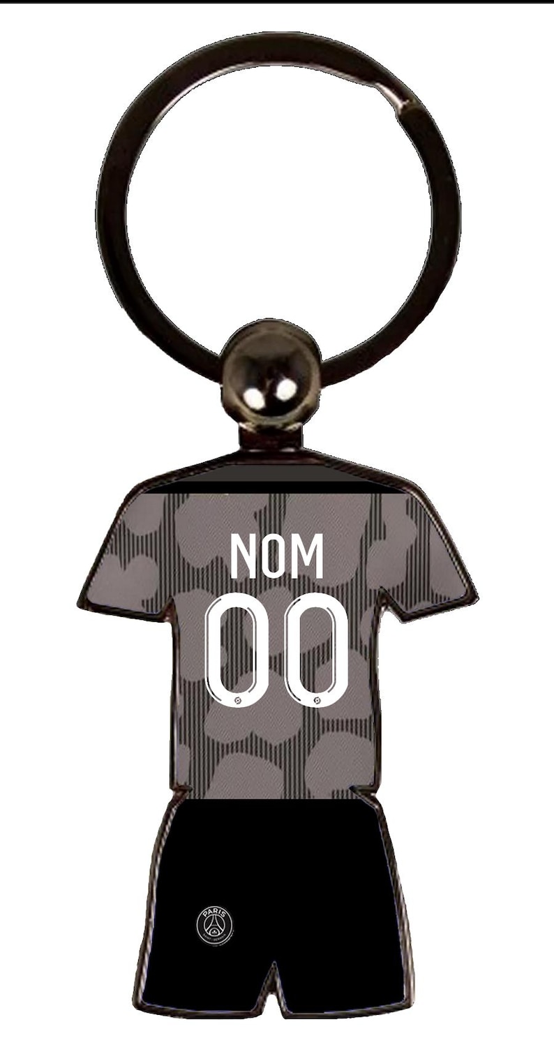 May include: A black and gray keychain in the shape of a soccer jersey and shorts. The jersey has the text "NOM" above the number "00". The keychain has a silver-colored ring and a small ball at the top.