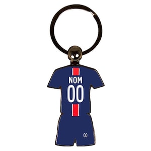 May include: A navy blue keychain in the shape of a soccer jersey with the number "00" in white. The jersey has red stripes and the word "NOM" in white. The keychain has a black metal ring.
