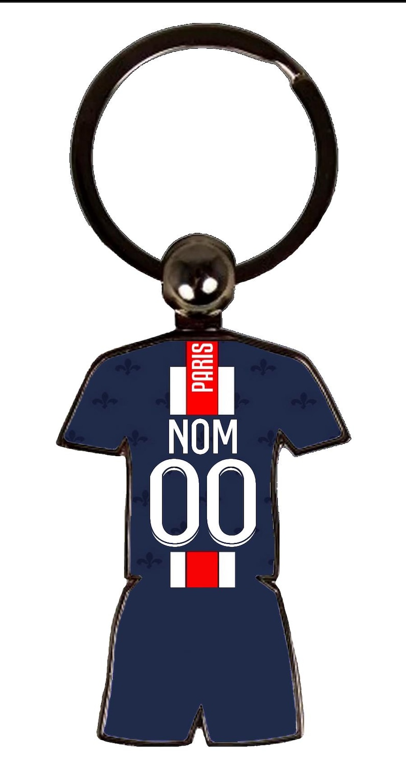 May include: A navy blue keychain in the shape of a soccer jersey. The jersey has the word "PARIS" in red, the word "NOM", and the number "00" in white. The keychain has a black ring.