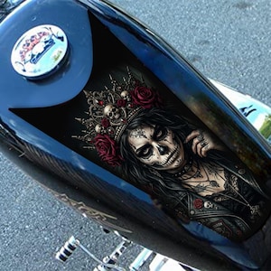 Könnte beinhalten: Schwarzer Motorradtank mit einem Grafikdesign einer Frau vom Tag der Toten, die einen Kranz aus Totenköpfen und Rosen trägt. Das Kunstwerk ist detailliert mit schwarzen, weißen und roten Farben. Der Tank hat eine glänzende, reflektierende Oberfläche.