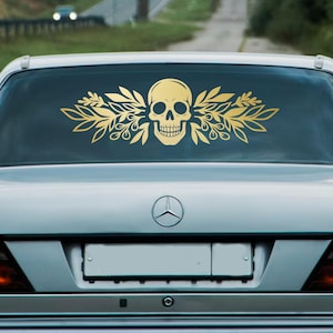 Op de afbeelding: Een goudkleurige schedel- en bladsticker is bevestigd op de achterruit van een zilveren auto. De schedel is gecentreerd, met bladeren die naar buiten uitsteken. De auto staat op een weg met een vage achtergrond.