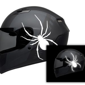 May include: A black motorcycle helmet with a white spider decal on the side. The helmet has a dark visor and a glossy finish. A smaller image shows the spider glowing.