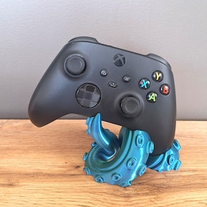 Könnte beinhalten: Ein mattschwarzer kabelloser Videospiel-Controller mit einem kreisförmigen Steuerkreuz, zwei Analogsticks und vier farbigen Aktionstasten mit der Beschriftung A, B, X und Y, der auf einem metallisch türkisfarbenen Krakenarm-Ständer ruht.