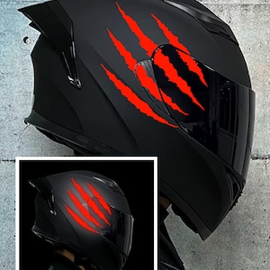 May include: Black motorcycle helmet with a red claw mark design. The helmet has a dark visor and a sleek, aerodynamic shape. The claw marks are a vibrant red, contrasting against the matte black finish. A smaller inset image shows the helmet with the claw marks glowing.