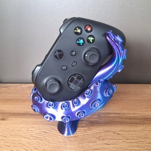 Könnte beinhalten: Ein schwarzer Xbox-Controller, der von einem 3D-gedruckten Ständer in Form eines gewundenen, violett-blauen Oktopus-Tentakels gehalten wird. Der Tentakel hat ausgeprägte Saugnäpfe und eine metallische Oberfläche, die den Controller sicher hält.