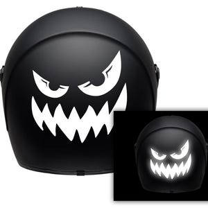 Op de afbeelding: Een zwarte motorhelm met een witte sticker die in het donker gloeit van een eng gezicht met ogen en tanden. De sticker is ontworpen om eruit te zien als een monster of een geest.
