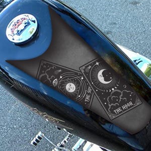 Könnte beinhalten: Schwarzer Motorradtank mit einem schwarzen Tankschutz mit zwei Tarotkarten-Designs. Eine Karte lautet "Die Sonne" und die andere "Der Mond". Die Karten haben himmlische Illustrationen in Weiß.