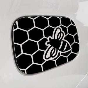 Könnte beinhalten: Schwarz-weißer Aufkleber mit Wabenmuster und einem Bienengrafik. Der Aufkleber ist auf einer weißen Oberfläche angebracht, wahrscheinlich der Tankklappe eines Fahrzeugs. Die Biene ist weiß umrandet und hat schwarze Streifen auf dem Hinterleib.