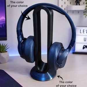 Pode incluir: Um suporte para auscultadores preto com um suporte vertical de forma oval e uma base circular. O suporte segura um par de auscultadores azul-escuro. O texto na imagem diz: The color of your choice, apontando para os auscultadores e a base.