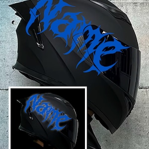 Puede incluir: Casco de moto negro con un diseño tribal azul. La palabra "Venge" está incorporada en el diseño. El casco tiene una visera oscura y una forma aerodinámica. El casco se muestra contra una pared de hormigón gris.