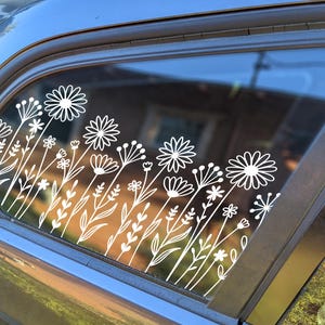 Puede incluir: Calcomanía de vinilo blanco de flores y hojas estilizadas en la ventana de un coche. La calcomanía es un dibujo de línea simple de una variedad de flores y hojas.