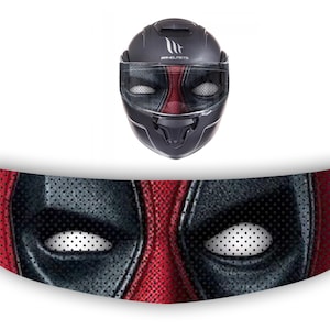 Puede incluir: Un casco de moto negro con un diseño de Deadpool rojo y negro. El casco tiene una visera transparente y una correa de barbilla negra.