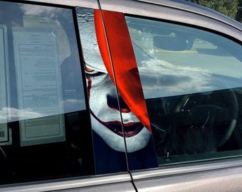 Op de afbeelding: Een autoruit met een vinyl sticker van een clownsgezicht met rode en witte make-up. Het gezicht van de clown is gedeeltelijk bedekt door een rood gordijn.