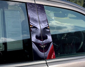 Op de afbeelding: Een auto raam sticker met een close-up van een clown gezicht met een sinistere glimlach. De ogen van de clown zijn felgeel en de sticker is gedrukt op een transparant vinyl materiaal.