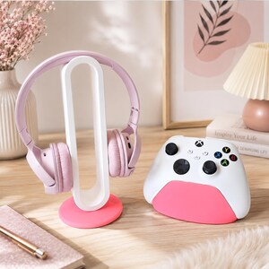 Op de afbeelding: Een lichtroze over-ear koptelefoon op een witte ovale standaard met een roze ronde voet, naast een witte en roze gamecontroller met zwarte joysticks en veelkleurige actieknoppen.