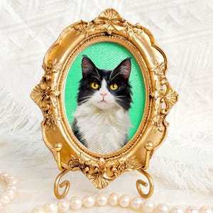 May include: A framed embroidered portrait of a black and white cat with yellow eyes. The oval portrait is set against a green background within an ornate gold-colored frame, displayed on a gold stand.
