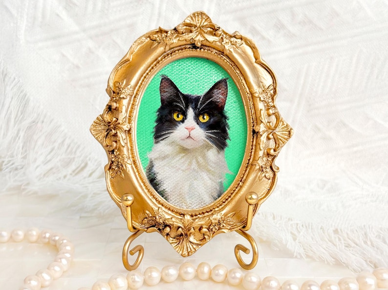 Acrylic portrait of a black and white long-haired cat in a vintage-style gold frame with a green background, surrounded by a pearl necklace.