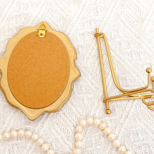 Back view of a vintage-style vertical oval gold frame with a hanging hook, placed next to a matching gold display stand on a woven white cloth.