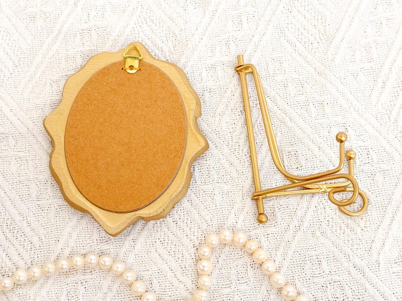 Back view of a vintage-style vertical oval gold frame with a hanging hook, placed next to a matching gold display stand on a woven white cloth.