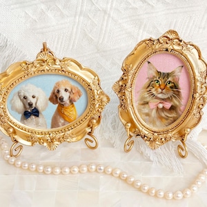 Two acrylic pet portraits in gold frames: one with a pair of white poodles in blue and yellow bandanas, the other with a Maine Coon cat wearing a pink bow