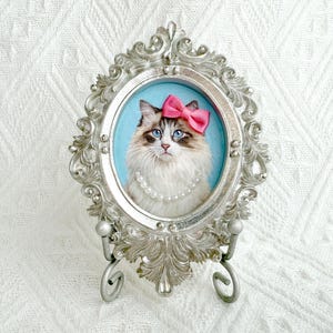 以下が含まれることがあります： 青い目、ピンクのリボン、真珠のネックレスをつけた猫の肖像画が、楕円形の装飾的な銀色のフレームに収められています。 猫は水色の背景に配置されています。 フレームは装飾的な銀色のスタンドで支えられています。