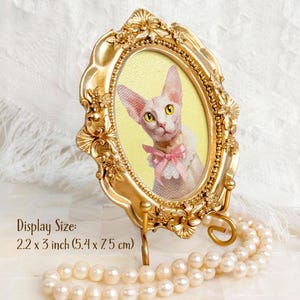 Portrait of a Sphynx cat in an ornate gold oval frame with yellow background and pink bow, displayed on a pearlescent surface with text showing display size: 2.2 x 3 inch (5.4 x 7.5 cm).