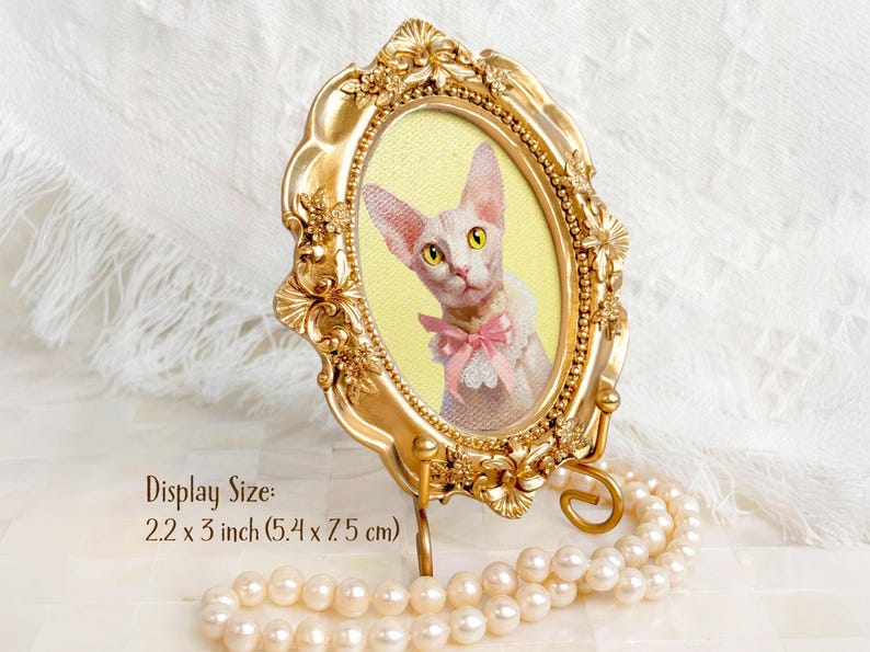 Portrait of a Sphynx cat in an ornate gold oval frame with yellow background and pink bow, displayed on a pearlescent surface with text showing display size: 2.2 x 3 inch (5.4 x 7.5 cm).